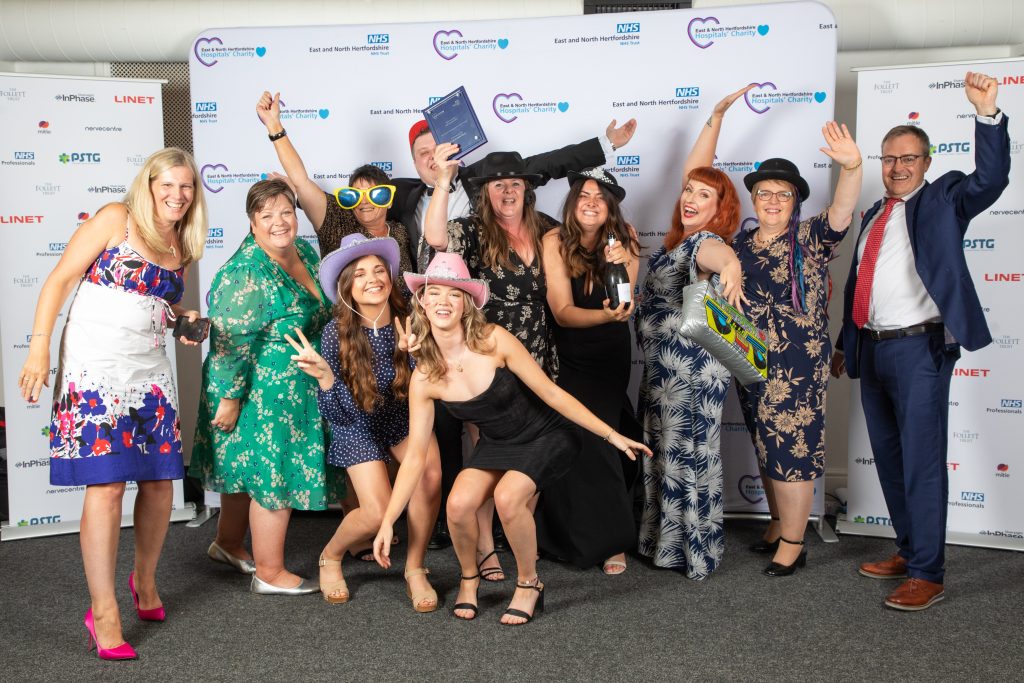 The critical care team wearing fancy dress items including cowboy hats, big sunglasses, police hats and holding a bottle of Champagne as they celebrate winning an award in front of a white backdrop with sponsors logos.