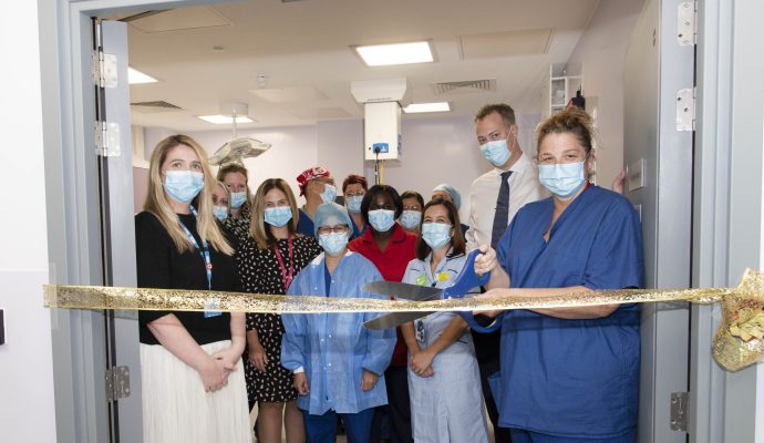 A member of staff in blue scrubs cuts a gold ribbon with big scissors in the doorway of one of the procedure rooms, as clinical and management staff watch on behind.