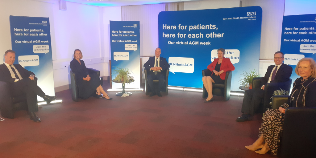 Members of the Trust's executive team sitting in socially-distanced seats in a semi-circle, with banners reading 'Here for patients, here for each other - our virtual AGM week' behind them.