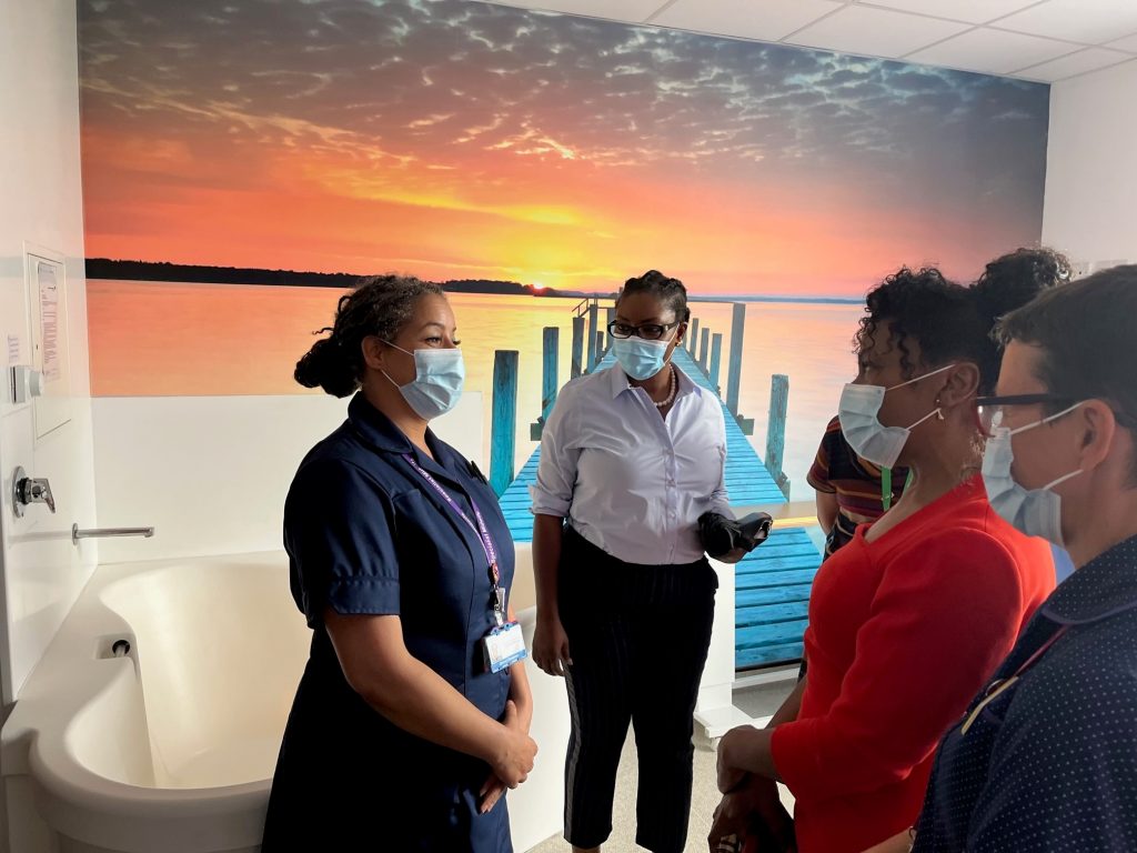 Midwife Dionne Thompson wearing her blue uniform and speaking to Professor Jacqueline Dunkley-Bent in a birthing pool room with a sea and sunset piece of art in the background and Wendy Olayiwola and Katie Chilton watching on.