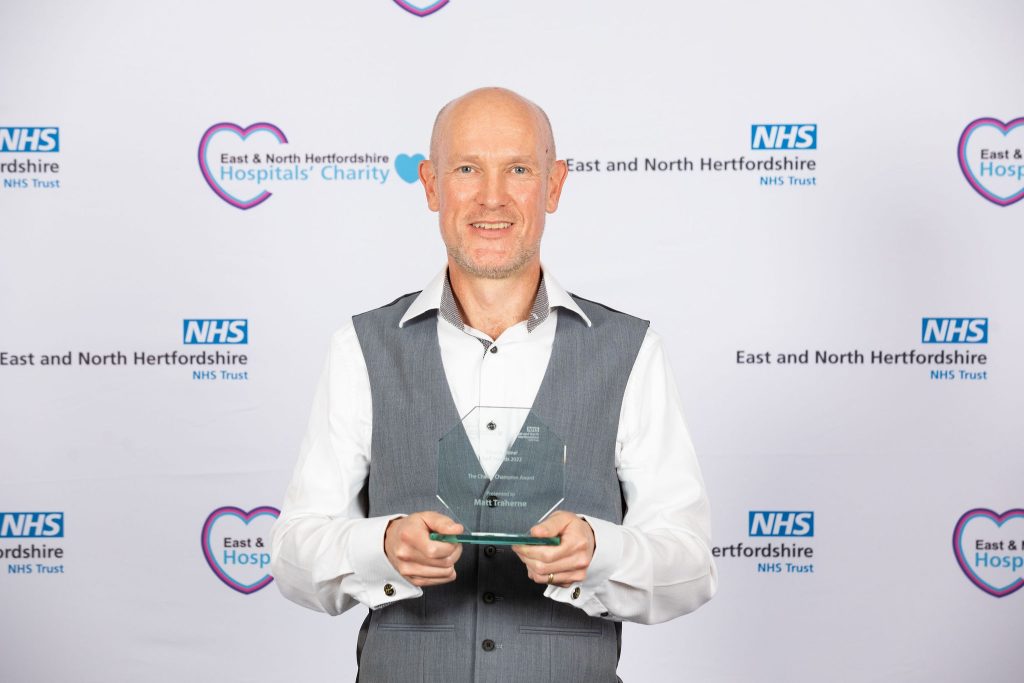 Matt Treherne smiling as he holds his glass award with both hands, in front of a white backdrop with sponsors logos.