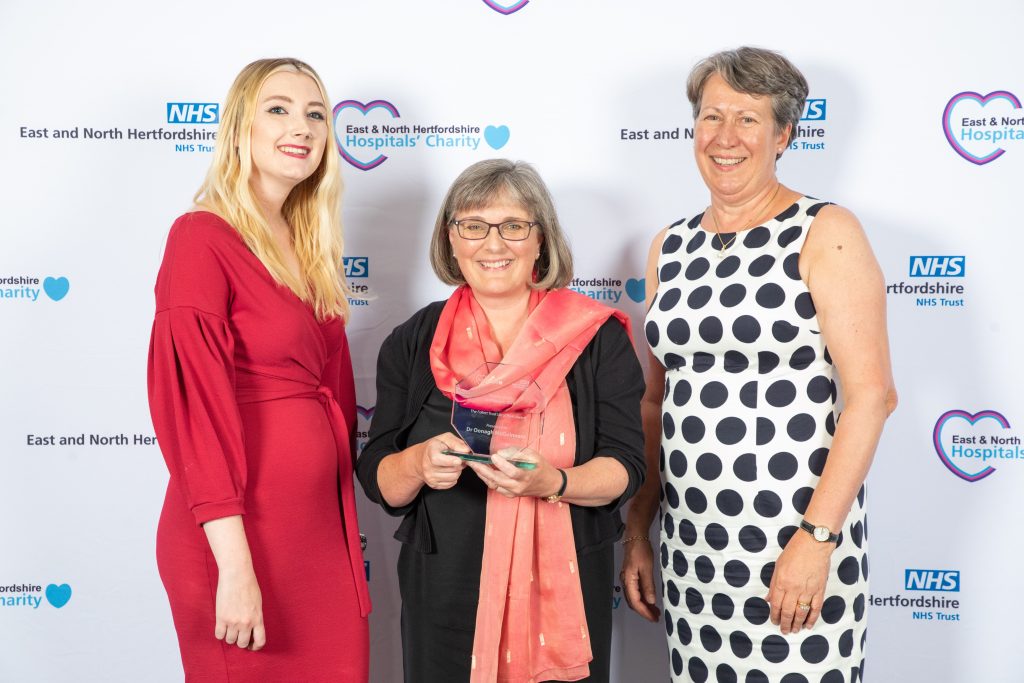 Dr Oonagh McGuinness pictured in the centre with her Follett Trust Local Hero Award alongside Comet editor Georgia Barrow (right) and Trust chair Ellen Schroder, in front of a white backdrop with sponsors logos.