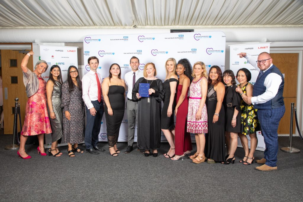 A smiling Covid Medicines Delivery Unit team with their award, against a white backdrop with sponsors logos.