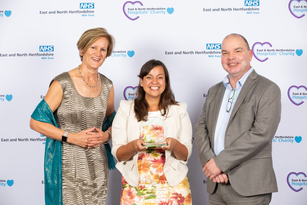 A smiling Claire Lyon (centre) picks up the award on behalf of Michelle Salisbury, in front of a white backdrop with sponsors logos.