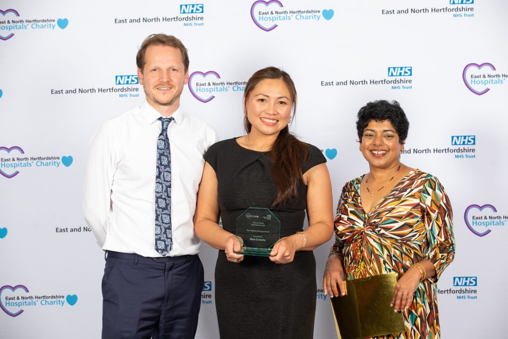 A smiling Mae Cometa with her award alongside Thom Pounds and Sylvia Gomez, against a white backdrop with sponsors logos.