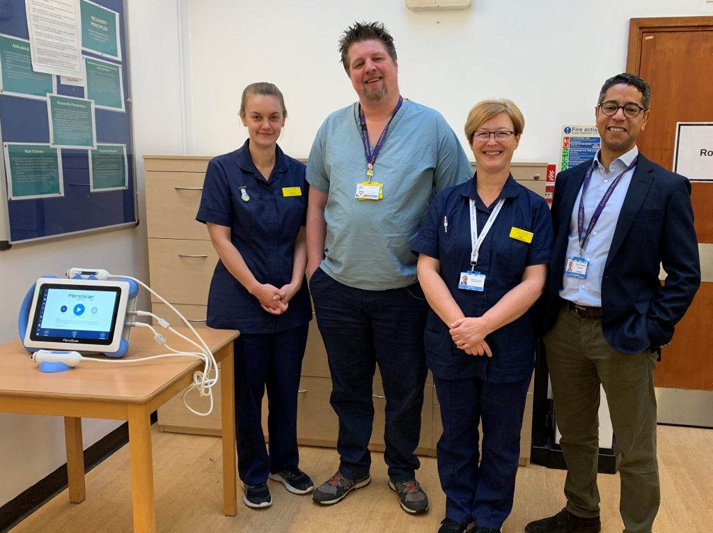 A FibroScan machine (looks like a digital tablet with wires coming out of it) on a table with smiling NHS staff in their uniforms next to it.