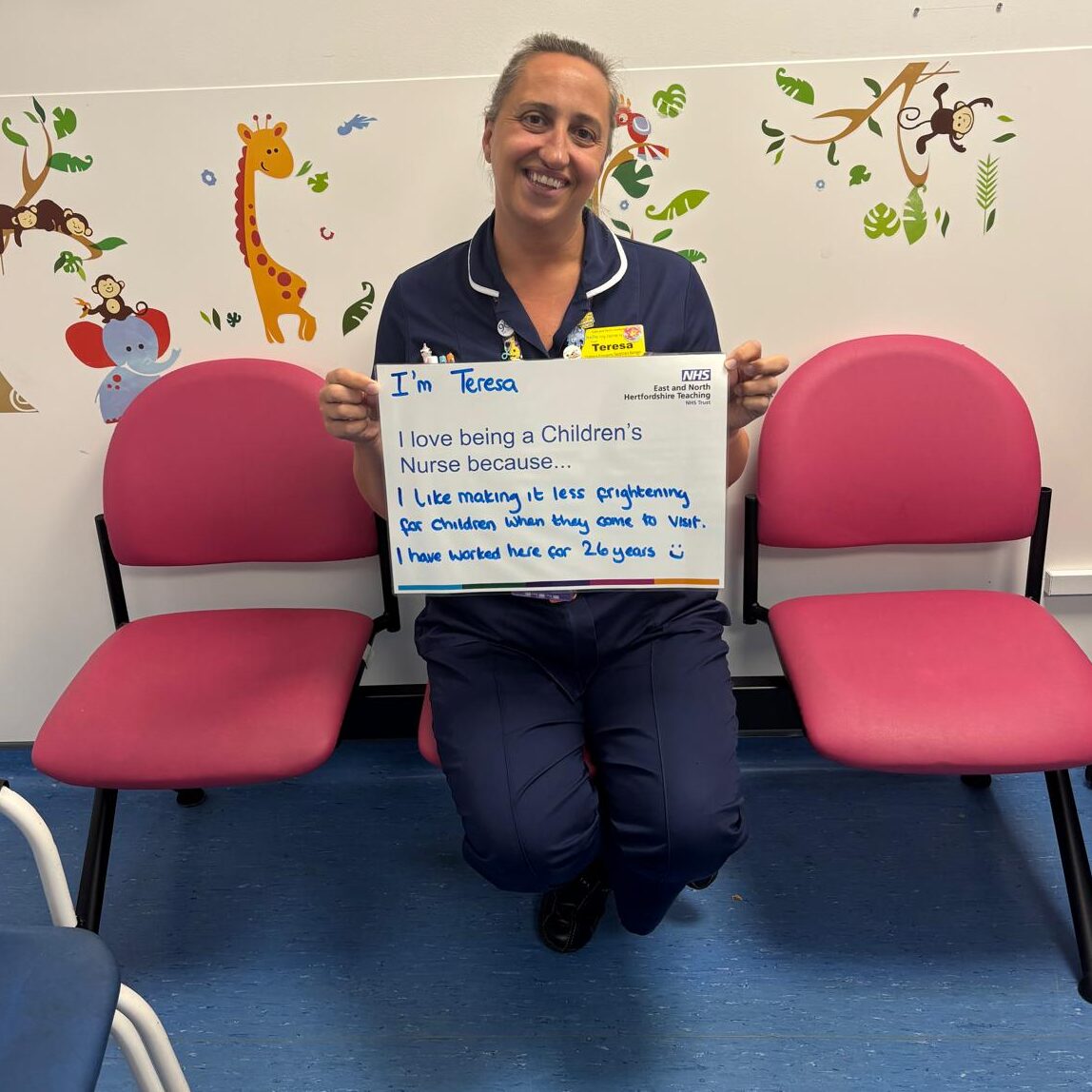 An image taken of a children's nurse. She is sat on pink chairs on an NHS ward holding a sign which details why she loves being a nurse.