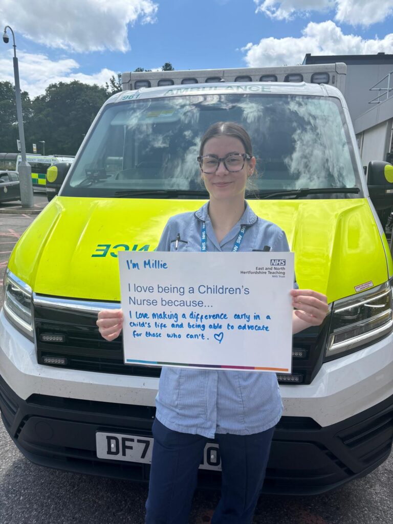 An image taken of a children's nurse. She is stood in front of an ambulance holding a sign which details why she loves being a nurse.