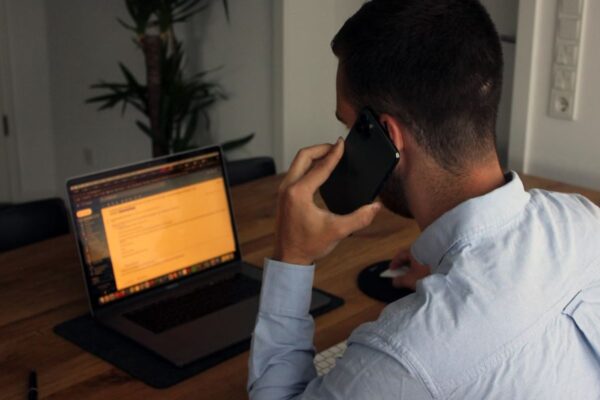 Man sat at a table with a laptop open, holding a mobile phone to his left ear.