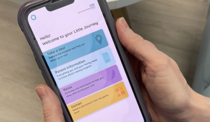 A photograph taken of the hands of one of the neonatal nurses at Lister Hospital. She is holding a mobile phone device which portrays the homepage of the Little Journey app.