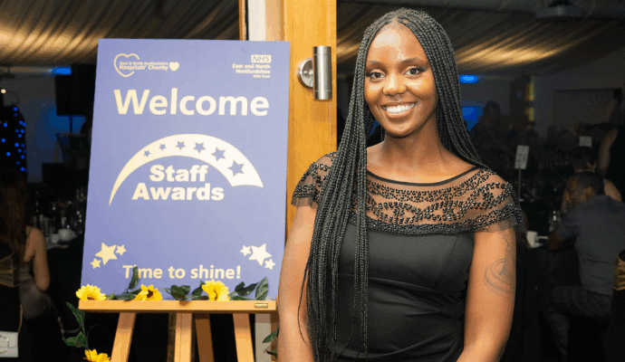A photograph taken during the Time to Shine Awards in 2024. A black woman is photographed smiling toward a camera. Behind her on an easel is a Staff Awards sign.