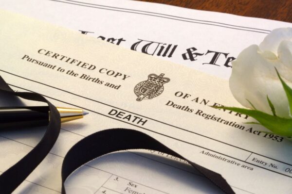 The top of a death certificate, with a black ribbon, a ballpoint pen, and part of a white rose resting on it.