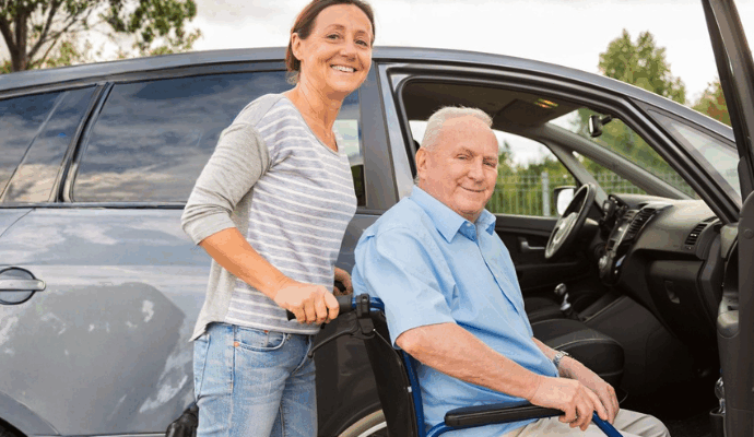 Photo of a female helping an older male wheelchair user into a vehicle.
