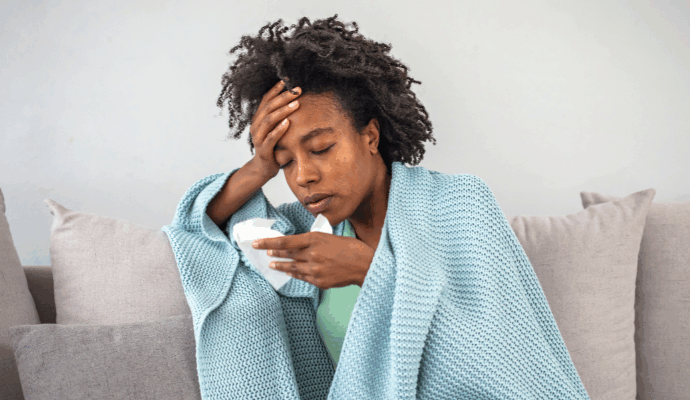 A photograph of a black woman sat on a sofa. She is draped in a warm blanket whilst visibly nursing flu symptoms.