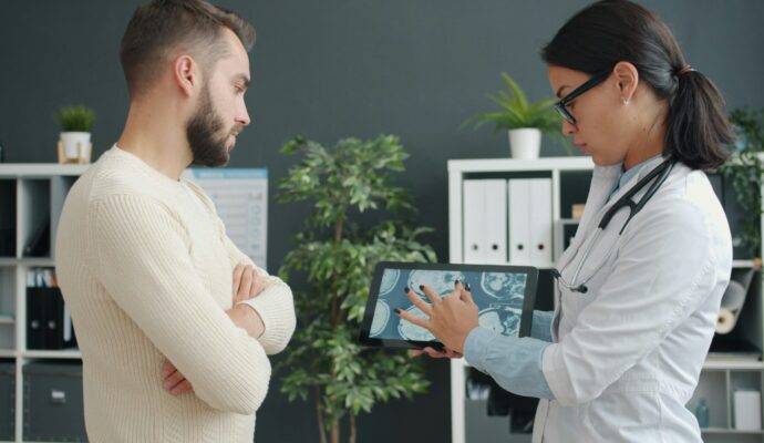 Female practitioner in uniform is showing male patient young man results on tablet screen in hospital office