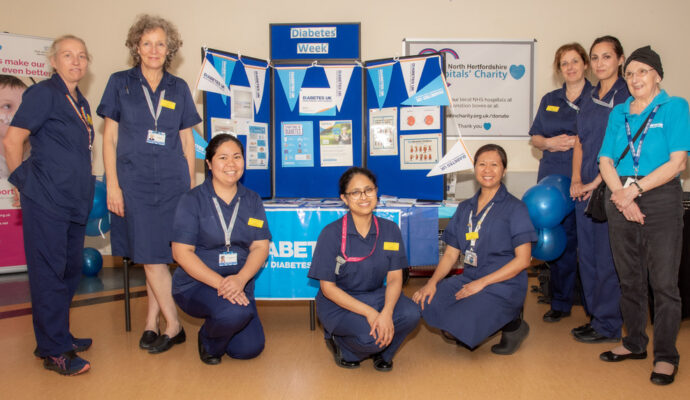 A picture taken during Diabetes Week of members of the diabetes nurse specialist team. The team are stood in front of an awareness stall with bunting and information posters. They are smiling toward the camera wearing nursing tunics.